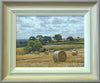 9 x 12 inch oil of round bales of straw on a hill sloping down from right to left, with distant trees and some nearer trees in the middle distance. Shows hand-finished frame, with gradated colours from off-white inner to beige/grey outer.