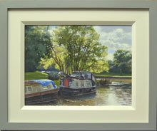 Load image into Gallery viewer, A 9 x 12 inch oil painting of the canal at GHillmorton, with a couple of narrowboats moored-up, with sunlight streaming through a tree on the far bank, with ripply reflections in the water, showing grey and cream frame
