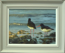 Load image into Gallery viewer, 12 x 16 inch Oil painting of a pair of Oystercatchers, paddling on the shoreline amongst stones, with light waves washing in, their red beaks and legs a colourful foil against the blue-green water, also showing the neutral grey frame
