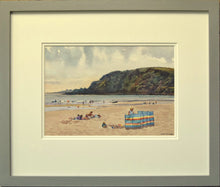Load image into Gallery viewer, Small watercolour, 7 x 10 inches, of Pentewan Beach in Cornwall, with the dark headland and colourful windbreak in the mid-ground, with many people having fun on the sand and in the surf, showing the double-mount and grey outer frame moulding.