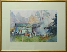 Load image into Gallery viewer, Two old sheds across the centre of the picture plane, with lots of paraphernalia stacked up against the left-hand shed, grass in the foreground and a pale backdrop of trees in the distance. Shows the cream mount and thin gold frame.
