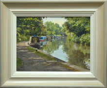 Load image into Gallery viewer, 9 x 12 inch oil painting of narrowboats moored next to the towpath on the left, with a bridge in the distance, trees reflected in the calm waters. Also shows frame with off-white slip, gradating to beige/grey outer frame.
