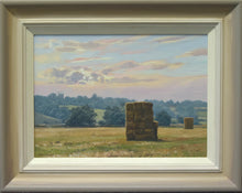 Load image into Gallery viewer, 10 x 14 inch oil painting of haystacks in a meadow, with trees in the background, ever bluer up to the horizon on the hill where Collyweston Church is on the left side of the skyline, with the sun breaking through a cloud. Also shows the frame with an off-white inner edge, gradating to a darker grey/beige outer edge.

