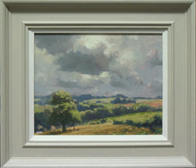 Load image into Gallery viewer, A view across the fields at Lyndon, with a big Oak tree in left foreground of stubble field, with distant blue trees and green fields and a lively, cloudy sky. Photo shows the grey frame with white inner slip.
