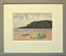 Load image into Gallery viewer, Small watercolour, 7 x 10 inches, of Pentewan Beach in Cornwall, with the dark headland and colourful windbreak in the mid-ground, with many people having fun on the sand and in the surf, showing the double-mount and grey outer frame moulding.
